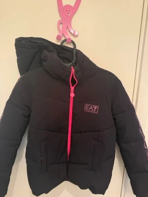 Svart pufferjacka EA7 med rosa detaljer - Snygg svart pufferjacka från EA7 Emporio Armani med rosa dragkedja och logga på bröstet. Jackan har huva och rosa tryck längs ärmarna. Perfekt för kalla dagar och ger en sportig look. Materialet är vadderat och håller värmen bra.