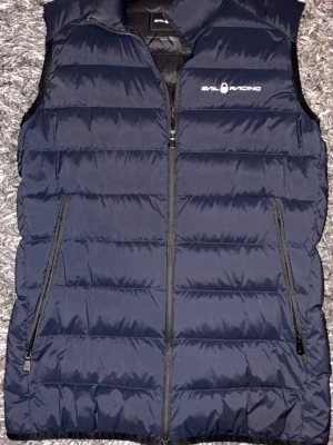 Marinblå dunväst Sail Racing - Snygg marinblå dunväst från Sail Racing med dragkedja framtill och två snedställda fickor med dragkedja. Västen har logga på bröstet och en detalj i nacken. Perfekt för lager-på-lager och håller dig varm med sin vadderade design.