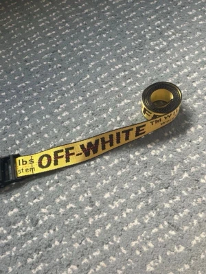 Gult Off-White bälte med text - Säljer ett ikoniskt gult bälte från Off-White med svart text och detaljer. Bältet har en industriell känsla med tydlig logga och svart spänne. Perfekt för att lyfta vilken outfit som helst och ger en streetwear vibe.