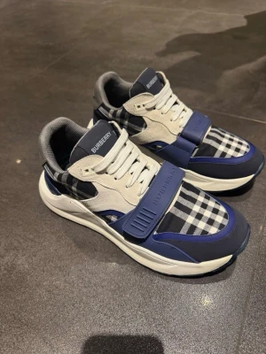 Burberry blåvita sneakers med rutmönster - Snygga sneakers från Burberry i blått och vitt med klassiskt rutmönster. Skorna har vita snören, blå detaljer och en bred rem över foten med Burberry-logga. Ovandelen är i textil och mockaliknande material, och sulan är vit och chunky.