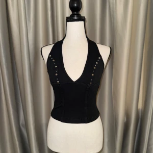 Y2K Svart korsett-topp med nitar - Snygg vintage korsett-topp i Y2K stil med djup v-ringning och dekorativa nitar längs axelbanden och framsidan. Toppen är figurnära och har breda axelband som ger en edgy look. Perfekt att styla med jeans eller kjol för en trendig outfit.
