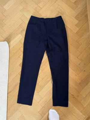 kostymbyxor J.Lindeberg  - Säljer ett par mörkblå kostymbyxor med rak passform och resår i midjan. Byxorna har knapp och gylf framtill samt två fickor bak med knapp. Perfekta för en clean och stilren look. Materialet känns mjukt och följsamt. Storlek 46. Nypris 1500 kr 