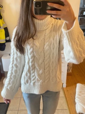 Vit kabelstickad tröja med polokrage - Säljer en vit stickad tröja med polokrage och klassiskt kabelstickat mönster. Tröjan har långa ärmar och breda ribbade muddar vid ärmslut och nederkant. Perfekt för att hålla sig varm under kyliga dagar.
