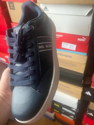 Marinblå sneakers från Björn Borg - Snygga marinblå sneakers från Björn Borg med vita detaljer och klassisk låg profil. Skorna har ovandel i slätt syntetmaterial, matchande blå snörning och vita ränder med logga på sidan. Perfekta för en stilren och sportig look.