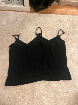 Svart linnetopp med smala axelband - Två svarta linnetoppar med smala axelband och enkel, stilren design. Perfekta att matcha med jeans eller kjol för en clean look. Tidlös modell som funkar till många olika stilar.