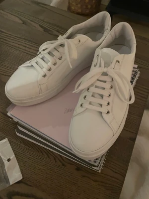 Vita Philipp Plein sneakers - Stilrena vita sneakers från Philipp Plein med klassisk låg profil och vita skosnören. Skorna har silverfärgade metalldetaljer och logotypen i silver bak på hälen. Tillverkade i slätt läder för en clean och modern look.