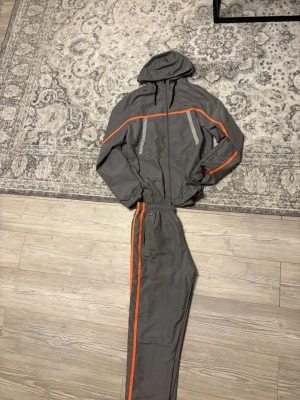 Grå träningsset med orange detaljer - Snyggt grått träningsset med matchande jacka och byxor. Setet har orange ränder längs ärmar och ben samt dragkedjor på jackan. Jackan har huva och resår i midja och ärmslut, byxorna har resår i midjan och vid benslut. Perfekt för träning eller chill.