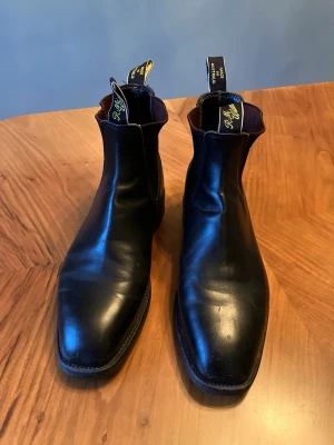 Svarta Chelsea boots från R.M. Williams - Säljer ett par klassiska svarta Chelsea boots från R.M. Williams i äkta läder. Boots med fyrkantig tå, elastiska sidopaneler och dragflikar med logga. Storlek 8 vilket motsvarar 42. De är inte stora i storleken, känns som mellan 7,5 och 8 (41,5-42). Lädersulan är sulad med en gummisula efter inköp för bättre motståndskraft mot regn. Mycket bra skick, inga skador. 