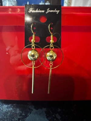 Guldiga statementörhängen med pärldetalj - Säljer ett par trendiga örhängen i guldfärgad metall De har en rund cirkel, en stor guldfärgad pärla i mitten och ett långt, smalt hängande stift. Perfekta för dig som gillar att sticka ut med accessoarer.