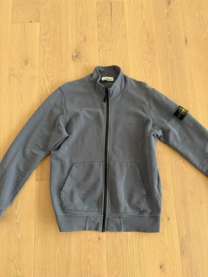 Blå zip hoodie från Stone Island - Säljer en stilren blå hoodie från Stone Island med dragkedja framtill och två stora fickor. Klassisk patch med logga på vänster ärm. Tillverkad i mjuk bomull, perfekt för en avslappnad och trendig look.