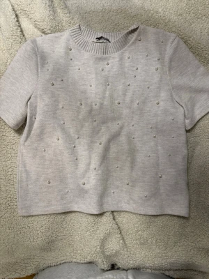 Ljusbeige topp med pärlor från Zara - Kortärmad ljusbeige topp från Zara med ribbad halsringning och dekorativa pärlor framtill. Toppen har en rak passform och är tillverkad i ett mjukt, stickat material som känns skönt mot huden. Perfekt för dig som gillar subtila detaljer och en clean look. Strl M men skulle säga passar S oxå💗väldigt bra skick.