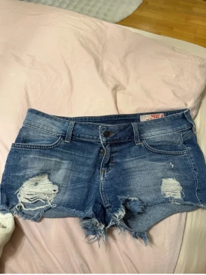 Blå slitna jeansshorts från Silver Jeans - Säljer ett par blå jeansshorts med slitna detaljer och fransig nederkant. Shortsen har broderade bakfickor och klassisk femficksdesign. Perfekta för sommarens chill dagar och har en cool, avslappnad look.