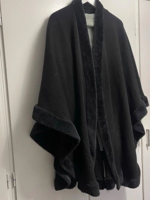 Svart ponchokappa med fuskpälsdetaljer - Stilren svart ponchokappa med breda ärmar och mjuka fuskpälsdetaljer längs kanterna och kragen. Kappan har en öppen front och ett avslappnat, svepande fall som ger en cool och mysig vibe. Perfekt för lager på lager och att slänga över vilken outfit som helst.