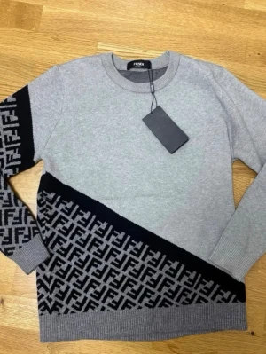 Grå stickad tröja från Fendi - Sny sopgrå stickad tröja från Fendi med svart diagonal panel och ikoniskt FF-mönster på ena ärmen och nedre delen. Rund halsringning och ribbade muddar. Perfekt för dig som gillar exklusiva detaljer och streetstyle.