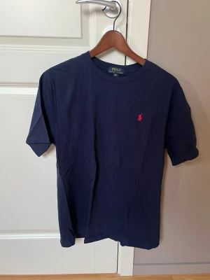 Mörkblå t-shirt från Polo Ralph Lauren - Klassisk mörkblå t-shirt från Polo Ralph Lauren med röd broderad logga på bröstet. T-shirten har rund halsringning och korta ärmar. Tillverkad i mjuk bomull som känns skön mot huden. Perfekt för en clean och stilren look.
