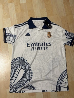  t-shirt Real Madrid Adidas drakmön ink - Säljer en vit Real Madrid t-shirt från Adidas med mörkblå detaljer och ett stort drakmönster. Tröjan har klubbmärke, Adidas-logga och ' sopor Fly Better' tryckt på bröstet. Tillverkad i lätt och ventilerande material, perfekt för fotboll eller träning.