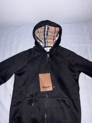 Svart — Burberry hoodie med dragkedja - Snygg svart hoodie från Burberry med dragkedja och klassiskt rutigt foder i huvan. Guldiga detaljer på snörena och dragkedjan ger en exklusiv känsla. Perfekt för dig som vill ha en stilren och lyxig look. Märkeslapp kvar på plagget. Säljer då den ej passa mig dessvärre, hade rekommenderat att vara max 175 för denna då den annars blir för kort.