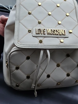 Beige ryggsäck från Love Moschino - Love Moschino ryggsäck – vit  Snygg och elegant ryggsäck från Love Moschino i vit färg med guldfärgade detaljer och nitar. Väskan är i mycket fint skick och använd väldigt lite.  Perfekt storlek för vardag – plats för mobil, plånbok, nycklar och andra små saker. Stilren design som passar till många outfits.  • Märke: Love Moschino • Färg: Vit med gulddetaljer   