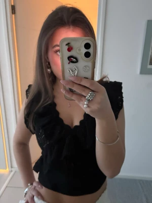 Svart spetsblus med volang - Säljer en svart spetsblus med v-ringning och volangdetaljer. Korta ärmar med ett vackert spetsmönster, vilket ger en romantisk touch! ❤️Köpt från zara. Säljer även samma i vit🥰