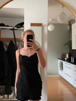 Svart satin klänning med smala axelband - Svart kortklänning i satin med smala axelband och enkel, stilren design. Klänningen har en lätt v-ringad hals och sitter tight över kroppen. 