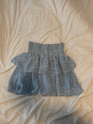 Blå volangkjol med blommigt mönster - Söt kort blå kjol med smockad midja och volanger. Kjolen har ett ljust blommigt mönster och är luftig, perfekt för varma dagar. Materialet känns lätt och bekvämt, och volangerna ger ett extra lyft till stilen.