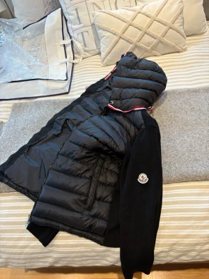 Svart pufferjacka från Moncler - Snygg svart pufferjacka från Moncler med stickade ärmar och huva. Jackan har dragkedja framtill, diskret Moncler-logga på ärmen och ribbad struktur på ärmarna. Perfekt för dig som vill ha en sportig och trendig look. Helt ny 