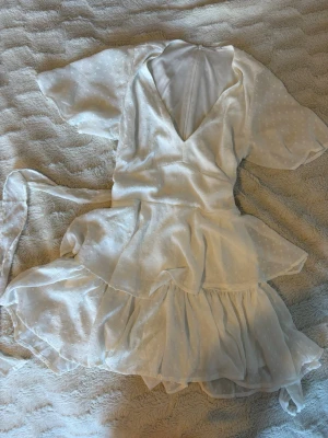 Vit kort klänning /studentklänning - Säljer en vit kort klänning med V-ringning. Klänningen har volangdetaljer samt inbyggda shorts och knyte baktill. Köpt från Vinted men tyvärr för stor för mig. Förra ägaren nämnde en liten defekt (se sista bilden), men jag har själv inte kunnat hitta den på klänningen.