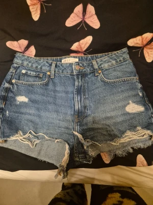 Blå denimshorts från Zara - Säljer ett par blå denimshorts från Zara med hög midja och råa, fransiga kanter. Shortsen har slitna detaljer framtill och bak, klassisk femficksdesign och knappgylf. Perfekta för sommaren och ger en avslappnad vibe.