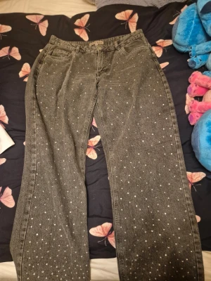 Svarta jeans med glitter - Säljer ett par svarta jeans med raka benupplägg och glitter över hela framsidan. Jeansen har klassisk femficksmodell och bälteshällor. Materialet är denim i bomull och de har en avslappnad passform som passar perfekt till en trendig vardagslook.