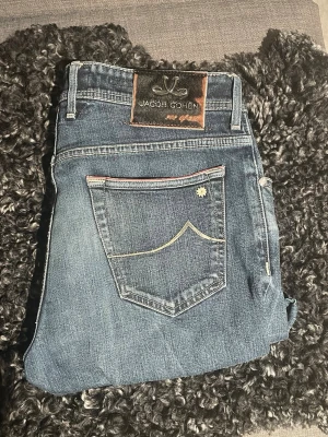 Blå jeans från Jacob Cohen - Snygga blå jeans från Jacob Cohen med klassisk femficksmodell och dekorativ söm på bakfickan. Jeansen har en läderpatch med logga bak i midjan och en liten blomdetalj på fickan. Perfekta för en avslappnad men stilren look.