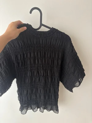 Svart croppad topp från Nelly - Trendig svart croppad topp från Nelly med smockad struktur och korta ärmar. Toppen har en lätt transparent känsla och vågig kant nertill för en cool look. Perfekt att styla med höga jeans eller kjol.