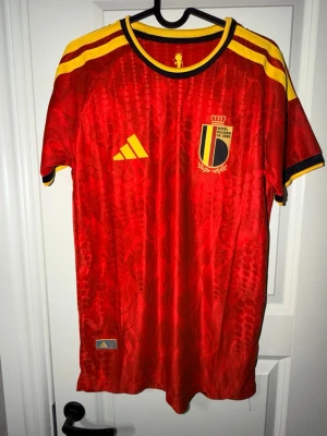 Belgien matchtröja Adidas röd - Säljer en röd Belgien matchtröja från Adidas med gula och svarta detaljer. Tröjan har korta ärmar, rund hals och Belgiska fotbollsförbundets emblem på bröstet. Tillverkad i lätt och ventilerande material, perfekt för fotbollsträning eller match. Sitter som small