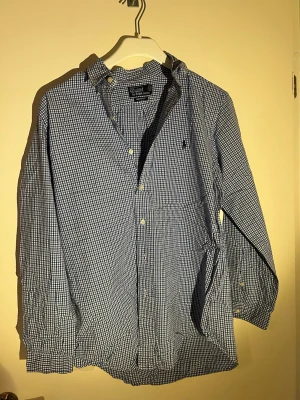 Blårutig skjorta från Polo Ralph Lauren - Klassisk blårutigu skjorta från Polo Ralph Lauren med button-down krage och broderad logga på bröstet. Skjortan har långa ärmar, vita knappar och är tillverkad i mjuk bomull. Perfekt för en avslappnad men ändå stilren look.