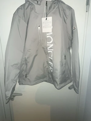 Grå vindjacka från Moncler - Snygg grå vindjacka från Moncler med huva och dragkedja framtill. Jackan har två sidofickor med dragkedja, justerbara ärmslut och Moncler-logga på ärmen. Stora vita Moncler-bokstäver på framsidan och diskret detalj i rött, vitt och blått på ryggen. Perfekt för blåsiga dagar.