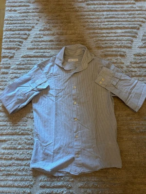 Blårandig skjorta från ZARA - Snygg skjorta från ZARA med tunna blå och vita ränder. Klassisk krage och knäppning framtill. Tillverkad i ett lätt bomullstyg som passar perfekt till sommaren. Avslappnad passform och ficka på ärmen för extra detalj.