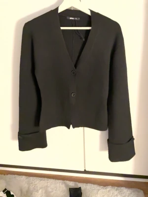 Svart kort kofta med knappar - Stilren svart kofta från GINA TRICOT med två knappar framtill och djup V ringning. Modellen är kortare och har långa ärmar med breda uppvik. Perfekt att slänga över en topp för en enkel men snygg look. Använd några fåtal gånger .