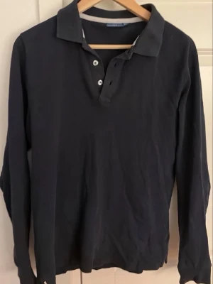 Mörkblå långärmad pikétröja - Klassisk mörkblå långärmad henley från Gant med tre knappar framtill och mjukt bomullsmaterial. Perfekt för en avslappnad och stilren look. Snygg krage och enkel design som passar till det mesta. Storlek M