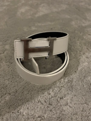 Svart-vitt bälte med silver H-spänne  - Snyggt vändbart bälte med stor H-spänne i metall. Ena sidan är vit och andra sidan svart, vilket gör det enkelt att liksom matcha till olika outfits. Bältet är gjort i slätt skinn och har en clean, modern look.