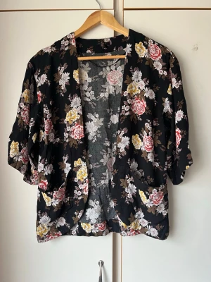 Svart blommig kimonos  - Säljer en svart kimonotopp med blommigt mönster i vitt, rosa och gult. Toppen har lös passform, trekvartsärm och öppen front. Perfekt för att slänga över ett linne eller klänning för en extra touch.