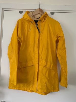 Gul Didriksons regnjacka med huva, S/M - Klassisk gul Didriksons 1913 regnjacka / regnkappa i storlek S/36. (Passar M också, men då med mer figurnära passform). Jackan har en huva med dragsko, dragkedja framtill med tryckknappar, samt två fickor med lock framtill. Jackan är designad för att vara 100% vattentät och har STORMsystem™-teknologi för att skydda mot väder och vind. Fint skick!