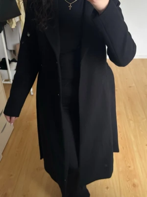 Svart lång kappa med knytskärp - Stilren svart lång kappa med klassisk krage och knytskärp i midjan. Kappan är tillverkad i ett mjukt tyg som ger en elegant look. 