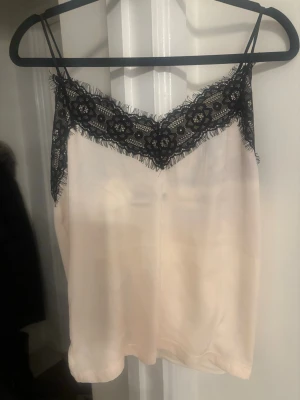 Beige satinlinne med spetsdetaljer - Snyggt beige linne i satin med tunna axelband och svart spets längs urringningen. Perfekt för att skapa en chic och trendig look. Materialet är glansigt och mjukt, vilket ger en lyxig känsla. Spetsen har ett blommigt mönster som ger linnet en extra touch.