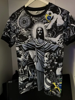 Svart t-shirt med Brasilien-tryck Nike - Unik svart t-shirt med färgstarkt och detaljerat tryck av Kristusstatyn och brasilianska motiv på framsidan. T-shirten har rund hals, korta ärmar och är tillverkad i ett mjukt syntetmaterial. Perfekt för dig som gillar iögonfallande och konstnärliga plagg.