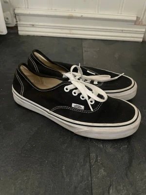 Svarta Vans Authentic sneakers - Klassiska svarta Vans Authentic sneakers med vit sula och vita snören. Skorna har låg profil, tydliga vita sömmar och Vans-logga på sidan samt röd 'Off The Wall'-logga bak. Ovandelen är i canvas och sulan i gummi.