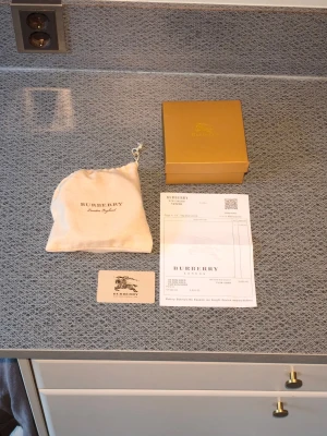 Burberry bälte med kvitto - Snyggt svart bälte från Burberry med klassiskt rutigt mönster på insidan och en stor silverfärgad 'TB'-logga som spänne. Bältet har exklusiv känsla och levereras med originalpåse box och kvitto..