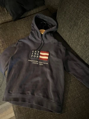 Mörkblå hoodie från Lexington - Snygg mörkblå hoodie från Lexington med broderad amerikansk flagga och texten 'American Heritage Since 1993' på bröstet. Klassisk huva med dragsko och mjukt material som känns skönt mot huden. Perfekt för en avslappnad och stilren look. I storlek 158-164