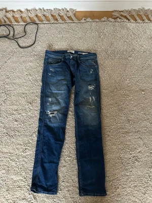 Blå jeans från Replay med slitningar - Säljer ett par blå jeans från Replay med slitningar och distressed detaljer på framsidan. Klassisk femficksmodell med raka ben och snyggt tvättad denim. Perfekt för dig som gillar en cool och avslappnad stil. Nytt skick på dem, inga defekter över huvudtaget. Storleken är W34 L34