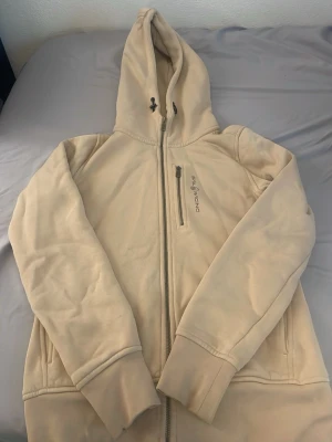 Beige hoodie från Sail Racing - Beige hoodie från Sail Racing med dragkedja framtill och en extra bröstficka med zip. Hoodien har justerbar huva med snören och ribbade muddar vid ärmslut och nederkant. Tillverkad i mjukt material som känns skönt mot huden.