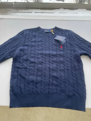 Blå kabelstickad tröja Polo Ralph Lauren - Kabelstickad blå tröja från Polo Ralph Lauren med rund halsringning och ribbade muddar. Tröjan har det klassiska röda Polo-logotypen broderad på bröstet och är tillverkad i mjukt material. Perfekt för dig som vill ha en stilren och tidlös look. Passar xl barn storlek och m i herrstorlek