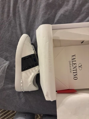 Vita Valentino Garavani sneakers - Snygga vita sneakers från Valentino Garavani med svart band och nitar över vristen. Skorna är i läder och har en stilren design med diskreta detaljer på sulan. Perfekta för dig som vill ha en lyxig och trendig look.